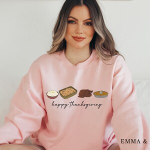 K&ouml;nnte beinhalten: Ein rosa Sweatshirt mit einer Grafik von Thanksgiving-Essen, darunter Kartoffelp&uuml;ree, F&uuml;llung, Truthahn und K&uuml;rbiskuchen. Der Text "happy Thanksgiving" ist unter der Essensgrafik gedruckt.