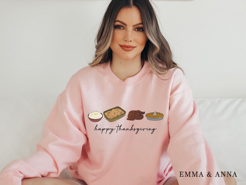 K&ouml;nnte beinhalten: Ein rosa Sweatshirt mit einer Grafik von Thanksgiving-Essen, darunter Kartoffelp&uuml;ree, F&uuml;llung, Truthahn und K&uuml;rbiskuchen. Der Text "happy Thanksgiving" ist unter der Essensgrafik gedruckt.