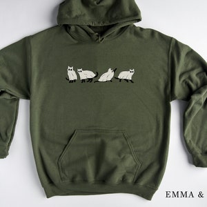 Puede incluir: Una sudadera con capucha verde con un gr&aacute;fico de cinco gatos blancos en la parte delantera. El texto "EMMA & ANNA" est&aacute; impreso en la esquina inferior derecha de la imagen.