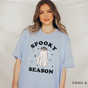 May include: A light blue t-shirt with a black graphic of a ghost wearing a flower crown. The text "Spooky Season" is printed in black below the ghost.