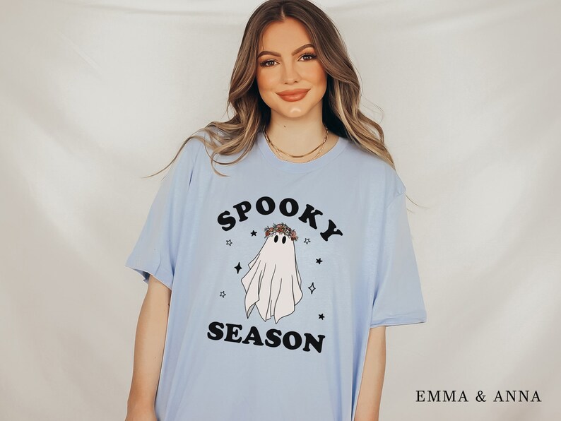 May include: A light blue t-shirt with a black graphic of a ghost wearing a flower crown. The text "Spooky Season" is printed in black below the ghost.
