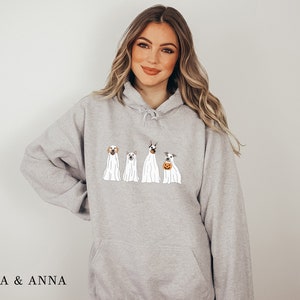 May include: A grey hoodie featuring a graphic of four dogs dressed as ghosts for Halloween. The dogs are wearing white sheets with their heads poking out. The text "EMMA & ANNA" is at the bottom of the image.