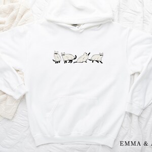 Puede incluir: Sudadera con capucha blanca con un gr&aacute;fico de cuatro gatos blancos con ojos y colas negros. Los gatos est&aacute;n en l&iacute;nea, caminando en la misma direcci&oacute;n.