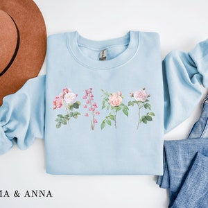 May include: A light blue sweatshirt with a floral print featuring pink and white roses and pink flowers. The sweatshirt is laid flat on a white surface with a brown hat and blue jeans.