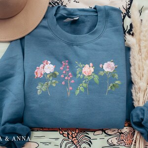 May include: A teal blue sweatshirt with a floral print of pink and white roses and pink flowers. The sweatshirt has a crew neck and long sleeves.