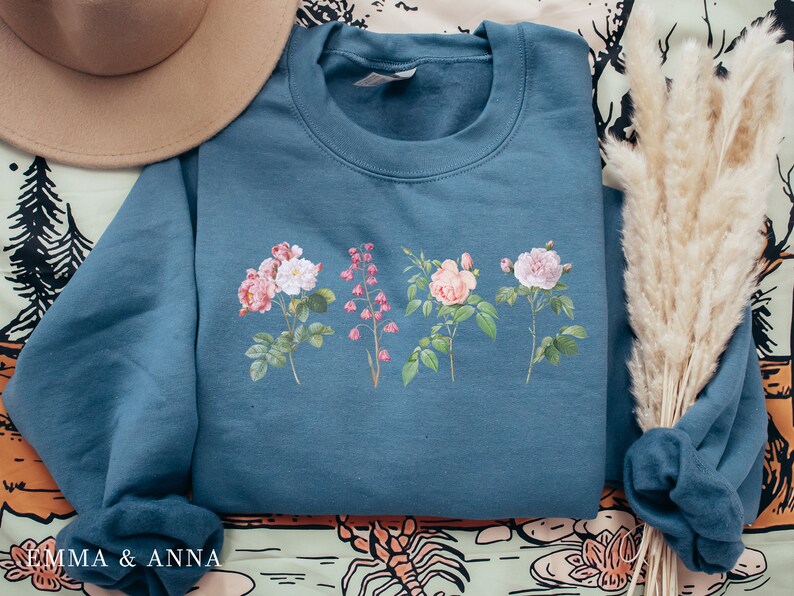 May include: A teal blue sweatshirt with a floral print of pink and white roses and pink flowers. The sweatshirt has a crew neck and long sleeves.