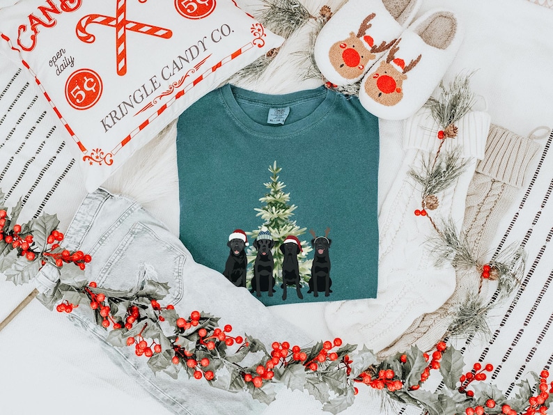 May include: A teal blue t-shirt with a Christmas tree and three black dogs wearing Santa hats. The dogs are standing in front of the tree. The t-shirt is surrounded by a white blanket, a red and white candy cane pillow, and a garland of holly.
