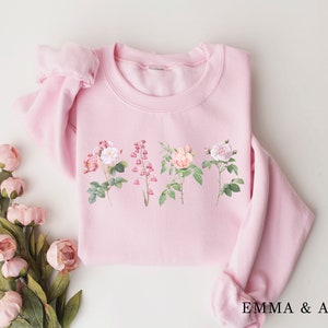 May include: A pink sweatshirt with a floral print featuring five different flowers. The flowers are pink, white, and green. The sweatshirt has a crew neck and long sleeves. The text "EMMA & ANNA" is printed on the bottom of the sweatshirt.