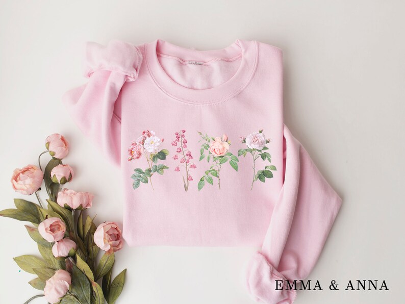 May include: A pink sweatshirt with a floral print featuring five different flowers. The flowers are pink, white, and green. The sweatshirt has a crew neck and long sleeves. The text "EMMA & ANNA" is printed on the bottom of the sweatshirt.