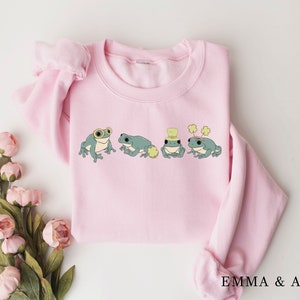Puede incluir: Una sudadera rosa con un gr&aacute;fico de cuatro ranas con sombreros y tr&eacute;boles. Las ranas son verdes con ojos negros y vientres blancos. La sudadera tiene cuello redondo y mangas largas.
