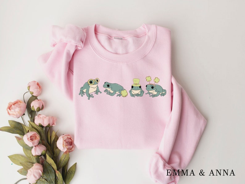 Puede incluir: Una sudadera rosa con un gr&aacute;fico de cuatro ranas con sombreros y tr&eacute;boles. Las ranas son verdes con ojos negros y vientres blancos. La sudadera tiene cuello redondo y mangas largas.
