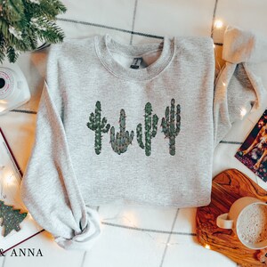 May include: A gray sweatshirt with a design of four green cacti with red and white lights on them. The sweatshirt is laying on a white and gray checkered surface with a book, a camera, a cup of coffee, and a Christmas card.