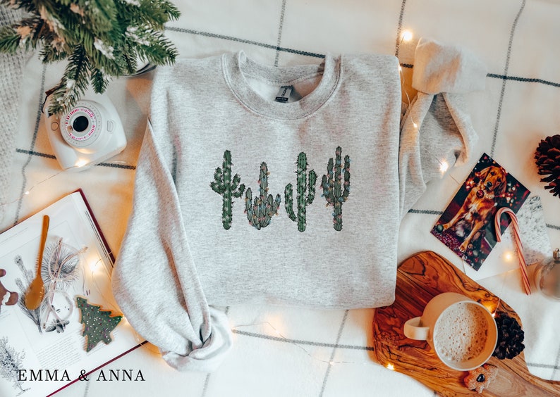 May include: A gray sweatshirt with a design of four green cacti with red and white lights on them. The sweatshirt is laying on a white and gray checkered surface with a book, a camera, a cup of coffee, and a Christmas card.