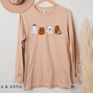 May include: A long-sleeved, light brown t-shirt with a graphic of four ghosts and pumpkins. The ghosts are wearing floral crowns and the pumpkins have stems and leaves.