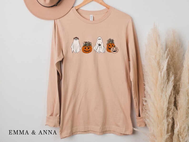 May include: A long-sleeved, light brown t-shirt with a graphic of four ghosts and pumpkins. The ghosts are wearing floral crowns and the pumpkins have stems and leaves.