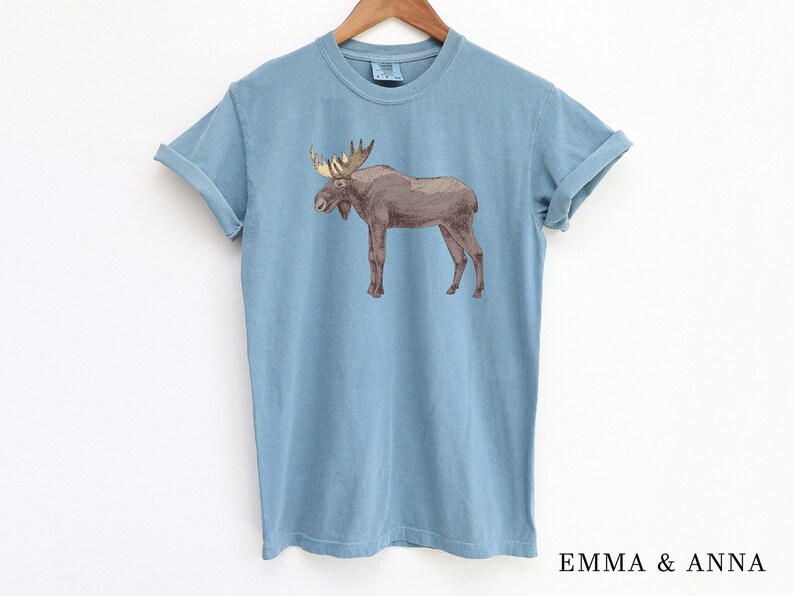 May include: A light blue t-shirt with a brown moose graphic. The moose has large antlers and is standing on all four legs.