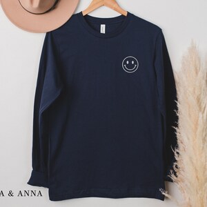 Puede incluir: Una camiseta de manga larga azul marino con una carita sonriente blanca en la parte delantera. La camiseta est&aacute; colgada en una percha de madera. El texto "EMMA & ANNA" est&aacute; en la parte inferior izquierda de la imagen.