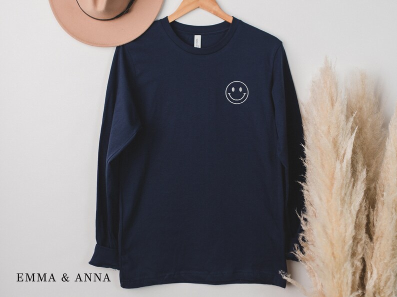 Puede incluir: Una camiseta de manga larga azul marino con una carita sonriente blanca en la parte delantera. La camiseta est&aacute; colgada en una percha de madera. El texto "EMMA & ANNA" est&aacute; en la parte inferior izquierda de la imagen.