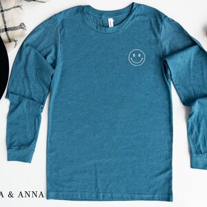 Puede incluir: Camiseta de manga larga color turquesa con una peque&ntilde;a carita sonriente bordada en el pecho. La camiseta est&aacute; hecha de un material suave y tiene cuello redondo. La marca "EMMA & ANNA" est&aacute; impresa en la parte inferior izquierda.