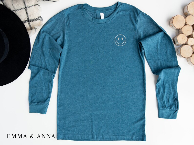 Puede incluir: Camiseta de manga larga color turquesa con una peque&ntilde;a carita sonriente bordada en el pecho. La camiseta est&aacute; hecha de un material suave y tiene cuello redondo. La marca "EMMA & ANNA" est&aacute; impresa en la parte inferior izquierda.