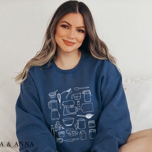 May include: A navy blue sweatshirt with a white graphic design of various kitchen tools and ingredients. The design includes a blender, a mixer, a rolling pin, a whisk, a measuring cup, a measuring spoon, a bowl, a jar, and a loaf of bread.