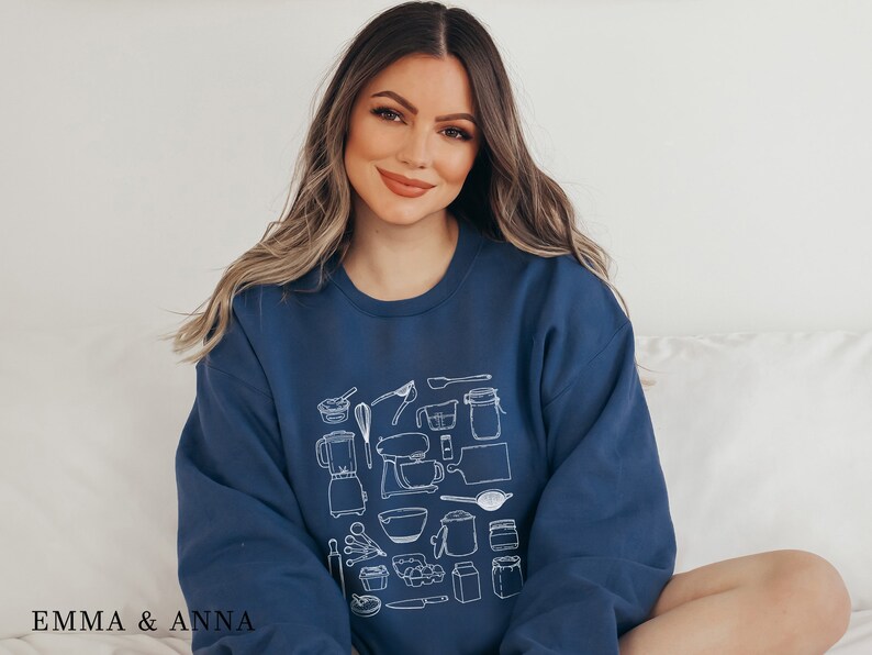 May include: A navy blue sweatshirt with a white graphic design of various kitchen tools and ingredients. The design includes a blender, a mixer, a rolling pin, a whisk, a measuring cup, a measuring spoon, a bowl, a jar, and a loaf of bread.