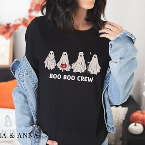 Puede incluir: Camiseta negra con un gr&aacute;fico de cuatro fantasmas vestidos como profesionales m&eacute;dicos. El texto "BOO BOO CREW" est&aacute; impreso debajo de los fantasmas.