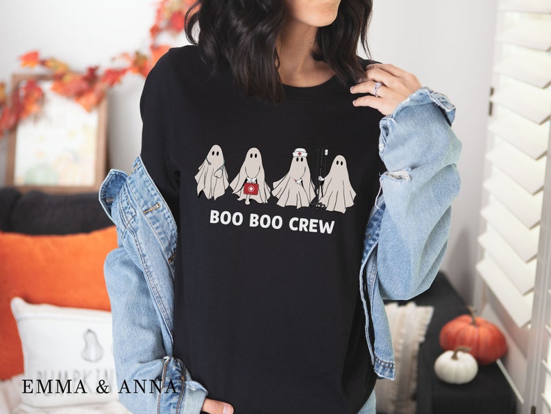 Puede incluir: Camiseta negra con un gr&aacute;fico de cuatro fantasmas vestidos como profesionales m&eacute;dicos. El texto "BOO BOO CREW" est&aacute; impreso debajo de los fantasmas.