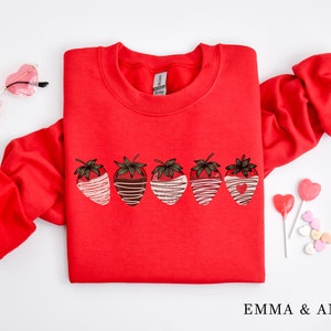 Puede incluir: Sudadera roja con un gr&aacute;fico de seis fresas cubiertas de chocolate. Las fresas est&aacute;n dispuestas en una fila y tienen rayas blancas y marrones. La sudadera tiene cuello redondo y mangas largas.