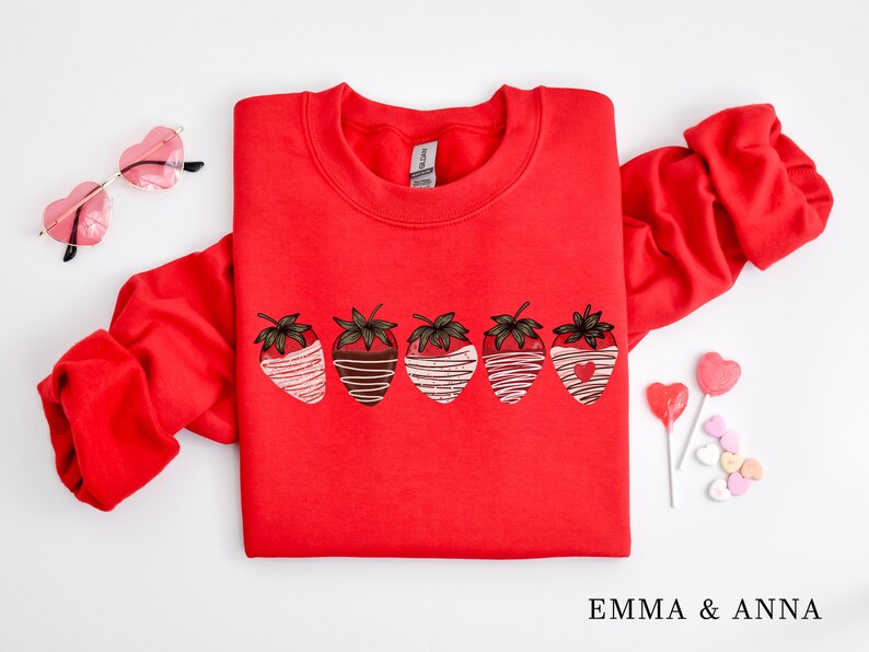 Puede incluir: Sudadera roja con un gr&aacute;fico de seis fresas cubiertas de chocolate. Las fresas est&aacute;n dispuestas en una fila y tienen rayas blancas y marrones. La sudadera tiene cuello redondo y mangas largas.