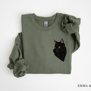 May include: A green sweatshirt with a black cat illustration on the front. The cat is looking to the left and has yellow eyes.