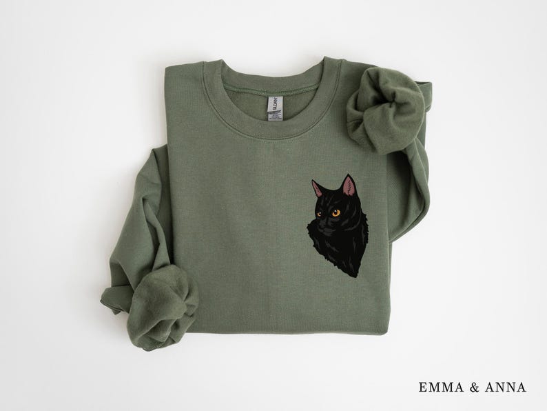 May include: A green sweatshirt with a black cat illustration on the front. The cat is looking to the left and has yellow eyes.