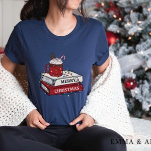 May include: A navy blue t-shirt with a graphic of a cup of hot chocolate with whipped cream and a candy cane, stacked on top of three books with the text "Merry Christmas" and Christmas lights.