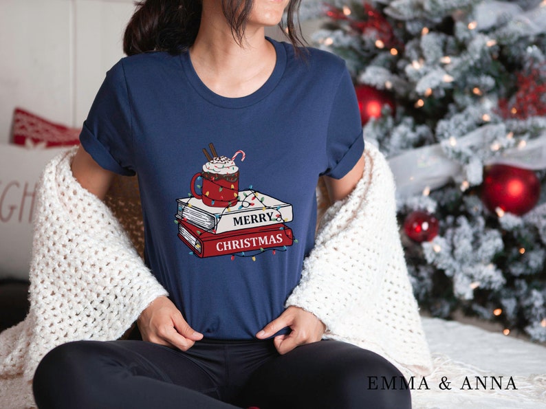 May include: A navy blue t-shirt with a graphic of a cup of hot chocolate with whipped cream and a candy cane, stacked on top of three books with the text "Merry Christmas" and Christmas lights.