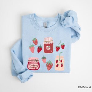 May include: Light blue sweatshirt with a strawberry jam theme. The sweatshirt features illustrations of jars of strawberry jam, strawberries, and a glass of strawberry juice. The text "Strawberry Jam" is printed on one of the jars.