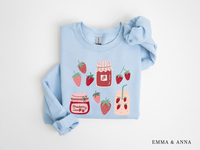 May include: Light blue sweatshirt with a strawberry jam theme. The sweatshirt features illustrations of jars of strawberry jam, strawberries, and a glass of strawberry juice. The text "Strawberry Jam" is printed on one of the jars.