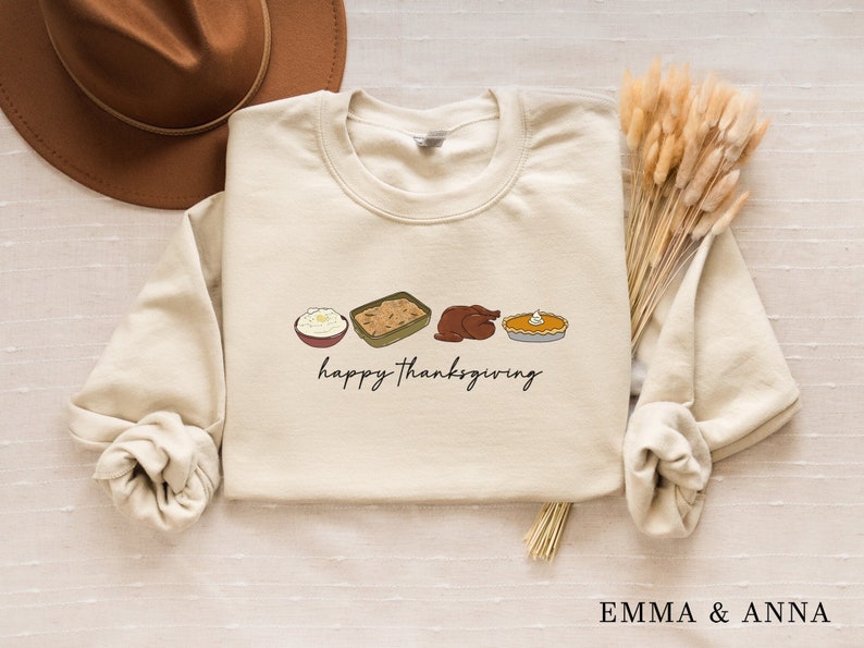 K&ouml;nnte beinhalten: Ein beiger Pullover mit dem Text "happy thanksgiving" und einer Reihe von Thanksgiving-Essen-Illustrationen: Kartoffelp&uuml;ree, F&uuml;llung, Truthahn und K&uuml;rbiskuchen.