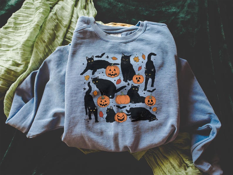 May include: A light blue sweatshirt featuring a Halloween-themed design. The graphic includes black cats, orange pumpkins with carved faces, and autumn leaves. The sweatshirt is folded, with a green fabric in the background.
