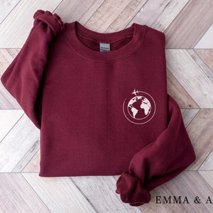 May include: Burgundy sweatshirt with a white graphic of a globe with a plane flying around it. The text "EMMA & ANNA" is printed on the bottom of the image.