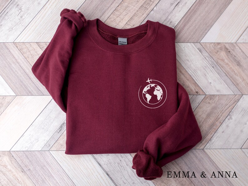 May include: Burgundy sweatshirt with a white graphic of a globe with a plane flying around it. The text "EMMA & ANNA" is printed on the bottom of the image.