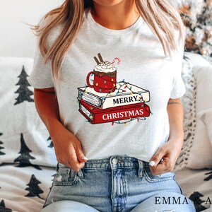 May include: White t-shirt with a graphic of a mug of hot chocolate with whipped cream and a candy cane on top of a stack of books with the words "Merry Christmas" and Christmas lights.