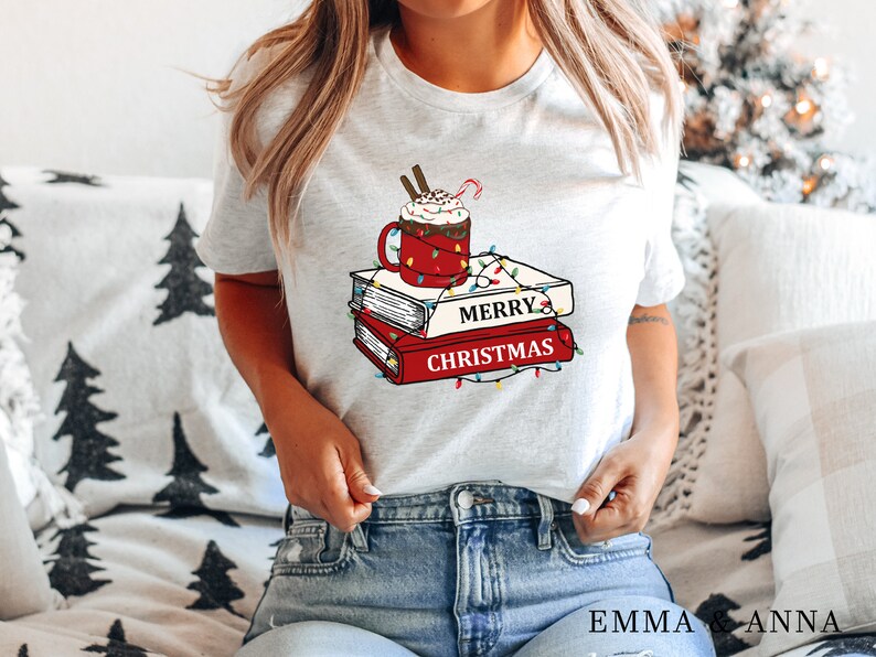 May include: White t-shirt with a graphic of a mug of hot chocolate with whipped cream and a candy cane on top of a stack of books with the words "Merry Christmas" and Christmas lights.
