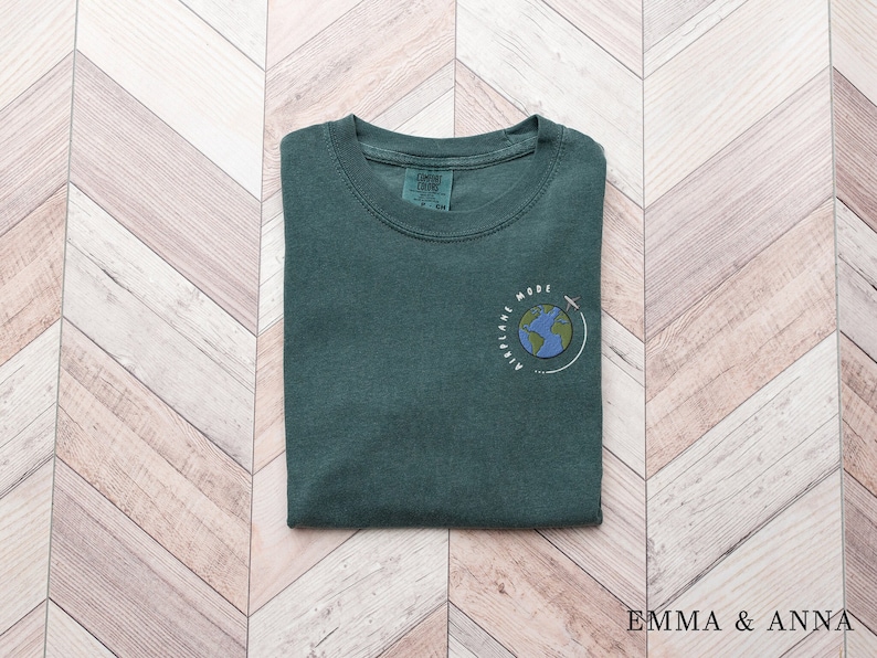 Op de afbeelding: Een donkergroen T-shirt met een wit geborduurd ontwerp van een wereldbol met een vliegtuig dat eromheen vliegt. De tekst "Airplane Mode" staat rond de wereldbol.