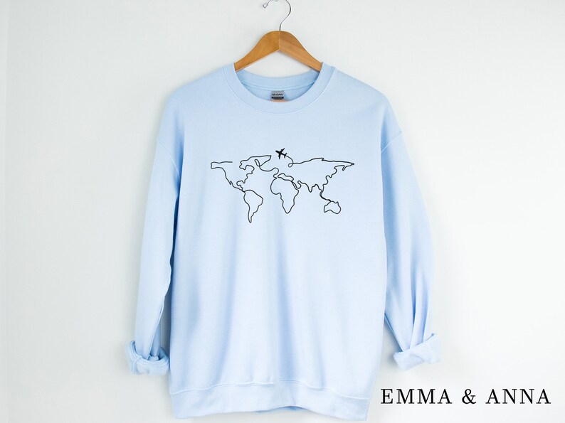 May include: A light blue sweatshirt with a black outline of a world map and a small airplane above it. The text "EMMA & ANNA" is printed on the bottom right corner of the sweatshirt.