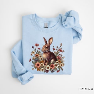 Pu&ograve; includere: Una felpa azzurra con un coniglio marrone circondato da fiori colorati. Il coniglio &egrave; raffigurato tra vari fiori. La felpa ha maniche lunghe. Il testo "EMMA & ANNA" &egrave; visibile in basso a destra.