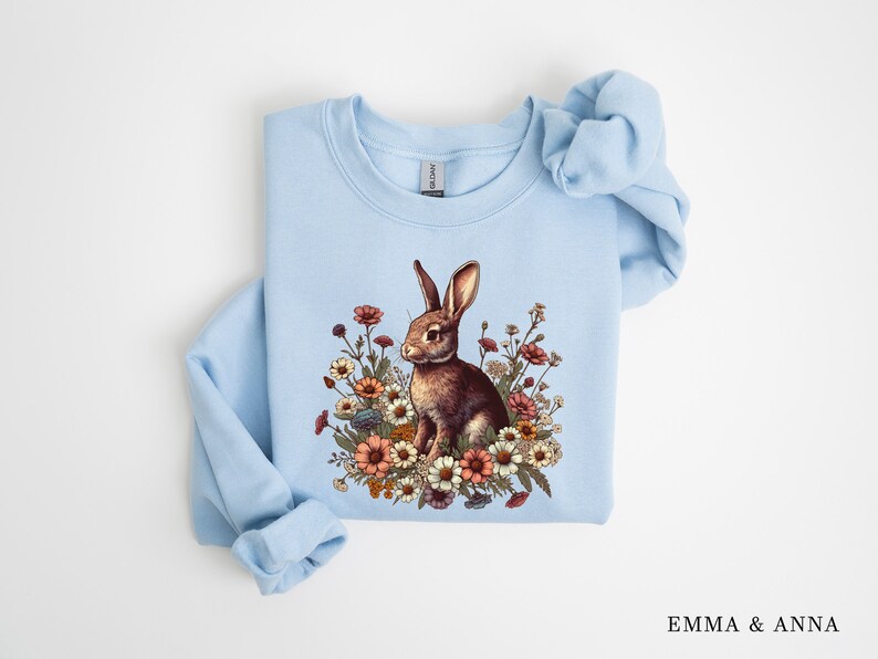 Pu&ograve; includere: Una felpa azzurra con un coniglio marrone circondato da fiori colorati. Il coniglio &egrave; raffigurato tra vari fiori. La felpa ha maniche lunghe. Il testo "EMMA & ANNA" &egrave; visibile in basso a destra.