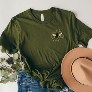 May include: A green t-shirt with a bee and honeycomb design embroidered on the front. The shirt is paired with blue jeans and a brown hat.