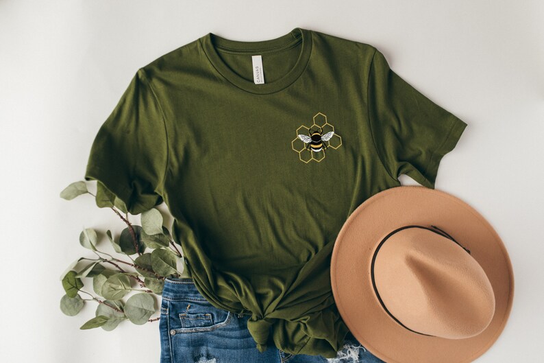 May include: A green t-shirt with a bee and honeycomb design embroidered on the front. The shirt is paired with blue jeans and a brown hat.