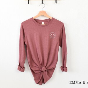 Puede incluir: Camiseta de manga larga color malva con un peque&ntilde;o smiley blanco en la parte superior de la espalda. La camiseta est&aacute; anudada a la cintura y colgada de una percha de madera. La marca "EMMA & ANNA" est&aacute; impresa en la parte inferior derecha.