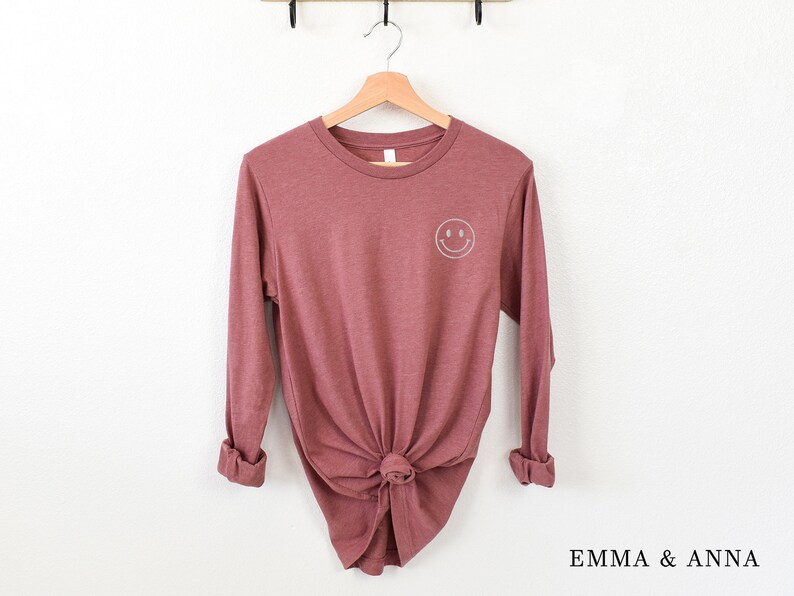 Puede incluir: Camiseta de manga larga color malva con un peque&ntilde;o smiley blanco en la parte superior de la espalda. La camiseta est&aacute; anudada a la cintura y colgada de una percha de madera. La marca "EMMA & ANNA" est&aacute; impresa en la parte inferior derecha.